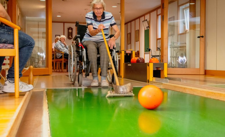 Dame spielt kegeln mit einer Bowlingkugel und einem Besen im Rollstuhl